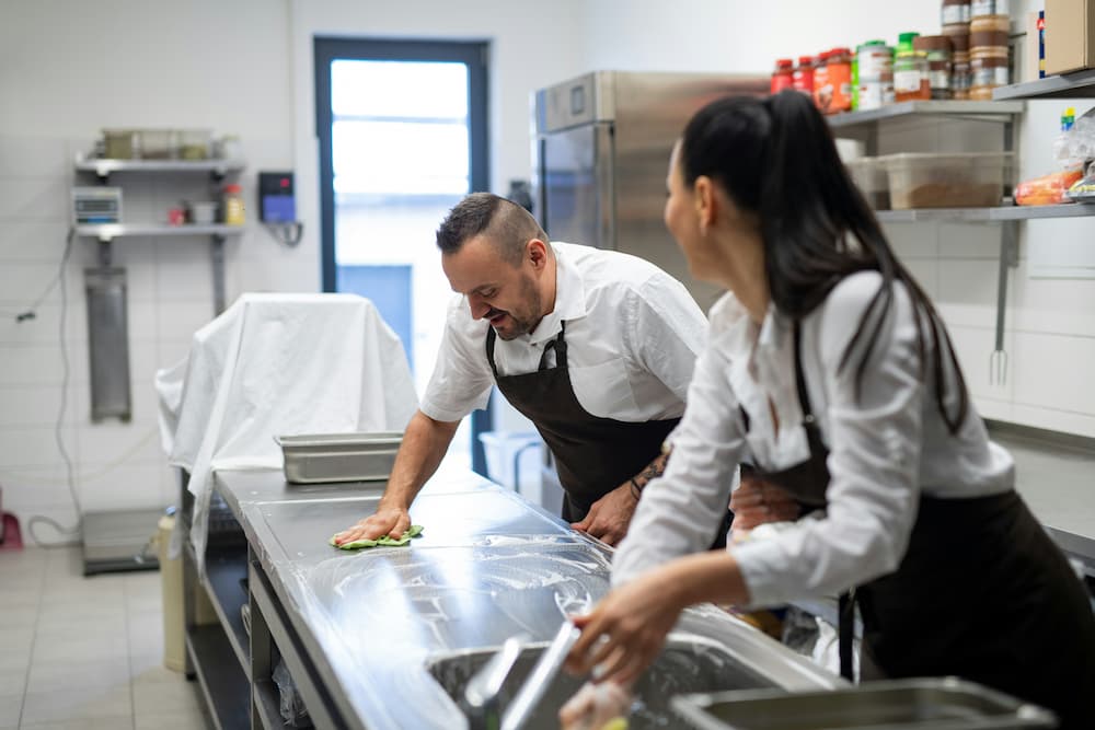 "Dois funcionários estão higienizando uma bancada de aço inoxidável em uma cozinha industrial. Ambos vestem uniformes brancos e aventais pretos, reforçando a importância de boas práticas de higiene no ambiente de trabalho.