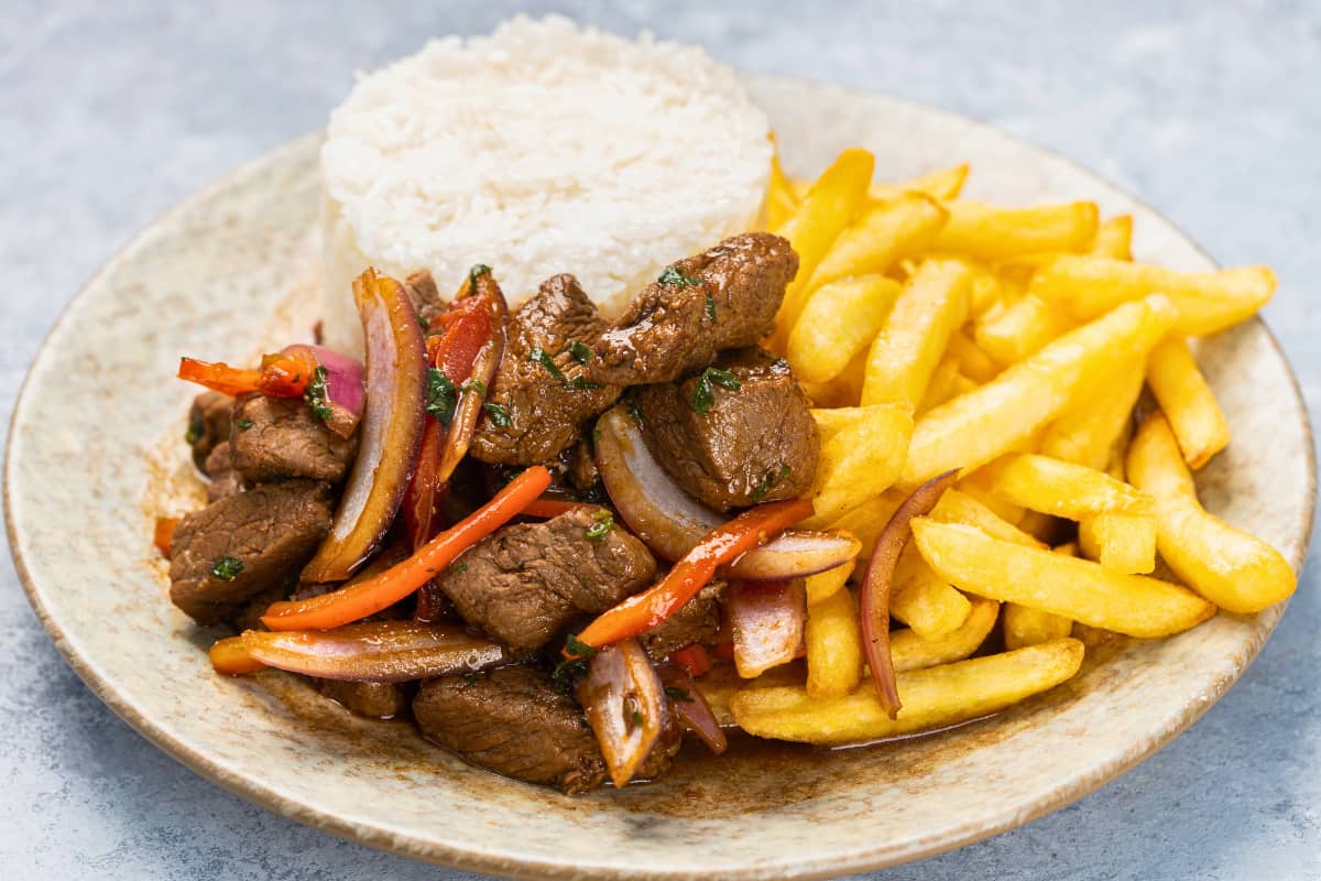 Prato Feito PF com carne, batata frita, arroz e vegetais coloridos em um prato rústico sobre uma mesa cinza.