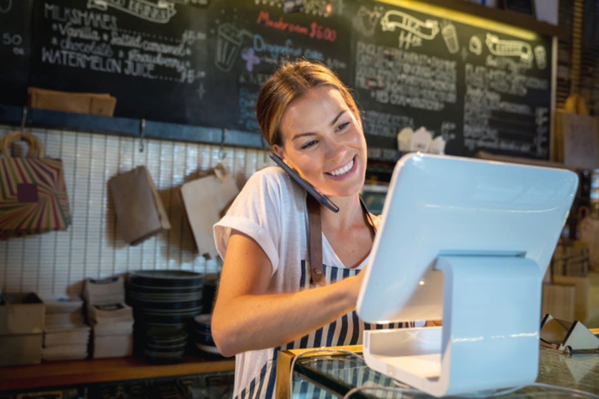 Pessoa sorridente trabalhando no caixa de um restaurante, respondendo clientes via celular.