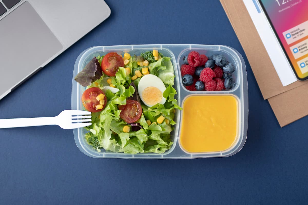 Imagem de marmita fitness com salada, ovo cozido, frutas vermelhas e molho, ao lado de um notebook, simbolizando como fazer marmita fitness para vender.