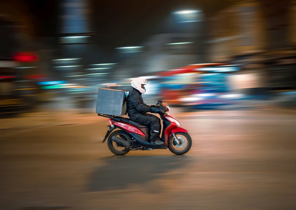 um entregador em uma motocicleta vermelha, usando capacete branco e jaqueta preta, percorre uma rua à noite com as luzes da cidade ao fundo