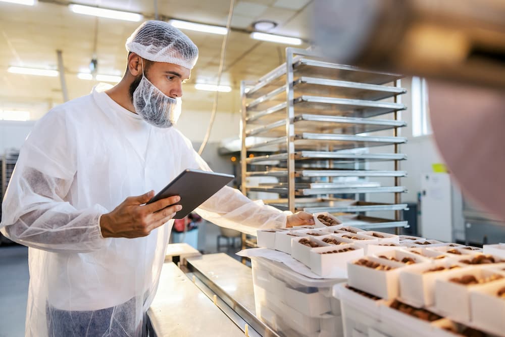 Profesional en una cocina profesional, utilizando una tableta y vistiendo ropa de protección, organizando productos.