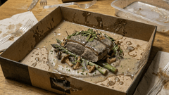 Um prato gourmet servido em uma caixa de apresentação escura sobre uma mesa de madeira clara. No centro, há um pedaço de carne coberto por um molho cremoso e ervas frescas, acompanhado de aspargos e cogumelos salteados.
