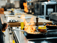 A imagem captura um momento dramático e dinâmico de cocção profissional em uma cozinha industrial, o que é comum em operações de alto volume como uma dark kitchen.