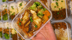 A imagem mostra uma marmita saudável sendo segurada por uma mão, em um recipiente plástico transparente. Dentro, há um filé de peixe grelhado com superfície dourada e levemente tostada, colocado sobre uma base de grãos (possivelmente arroz ou lentilhas).