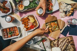 Una vistosa toma aérea (flat lay) de una mesa de madera pintada de rosa brillante, repleta de diversos platos de comida callejera internacional.