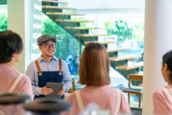 Chef sonriente en un café, usando un delantal y sosteniendo una tableta, conversando con el equipo en un ambiente moderno e iluminado.