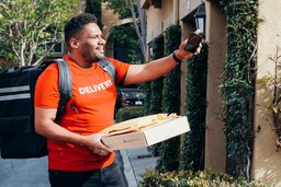 Homem segurando uma caixa e mochila de entrega preta, usando camiseta laranja "delivery" e calça jeans preta.