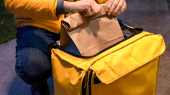 Esta imagen muestra a un repartidor de comida. El repartidor lleva una chaqueta amarilla de manga larga y pantalones vaqueros. Está agachado y sujetando una bolsa de papel marrón que contiene comida.