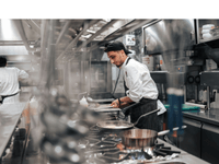 A imagem mostra o interior de uma cozinha industrial profissional. Em primeiro plano, um cozinheiro está trabalhando concentrado, usando uniforme branco com avental escuro e boné preto virado para trás.