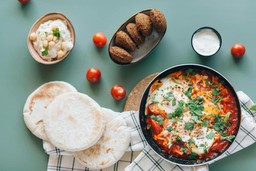 Uma comida árabe disposta em flat lay, apresentando falafel, hummus, pão pita e shakshuka, com tomates espalhados ao redor.