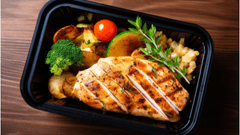 Uma marmita em embalagem preta contém um peito de frango grelhado fatiado, com marcas douradas de grelha. Ao redor, há acompanhamentos como brócolis, batatas assadas, tomate-cereja, uma rodela de limão grelhado e um ramo de ervas (alecrim).