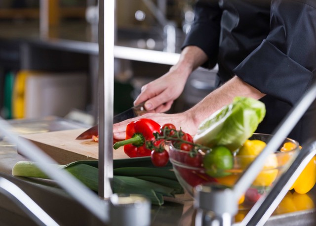 Sustentável: mãos masculinas cortando vegetais, homem está vestido com uniforme preto, em cima da bancada dele está vários legume e vegetais
