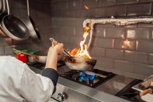 A imagem mostra uma cozinha profissional em funcionamento intenso, com um cozinheiro manuseando uma frigideira sobre uma chama alta de fogão industrial, gerando labaredas típicas de uma técnica conhecida como flambagem.