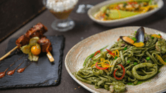 Presenta una mesa con platos típicos de la cocina peruana. En primer plano resalta un plato de tallarines verdes con mariscos (con conchas de abanico, calamares y langostinos). A la izquierda, sobre una fuente de pizarra negra, se sirven unos tradicionales anticuchos acompañados de un aguaymanto (physalis) y gotas de salsa.