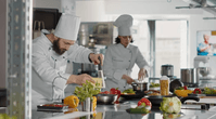 Dois chefs de cozinha cozinhando em uma cozinha para delivery. Tanto o homem quanto a mulher usam dólmã.