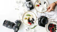 Uma vista superior artística (flat lay) sobre uma superfície branca, misturando comida e equipamentos fotográficos.