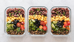 A imagem mostra uma vista superior (flat lay) de três recipientes de vidro retangulares perfeitamente alinhados, contendo refeições saudáveis e coloridas (meal prep), dispostos sobre uma superfície clara e levemente texturizada.