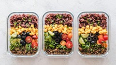 Três marmitas de vidro organizadas lado a lado, contendo refeições saudáveis e coloridas. Cada uma tem camadas de arroz integral ou arroz negro, grão-de-bico, lentilha, pepino, tomate-cereja, azeitonas e ervas.