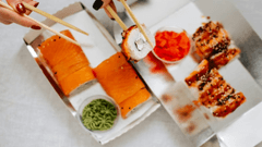 A imagem apresenta uma visão aérea em close-up de uma refeição de sushi para viagem, servida em embalagens de papelão prateadas sobre uma superfície clara.