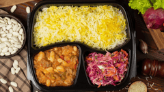 Uma marmita dividida em três compartimentos: arroz branco com açafrão na parte maior, feijão branco com carne em molho na lateral e uma salada colorida com repolho roxo e cenoura.