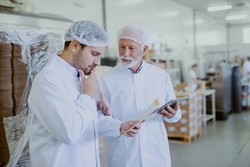 Dois funcionários de uma indústria alimentícia estão em um ambiente de produção, ambos usando jalecos brancos, toucas de proteção e luvas. Um deles segura uma prancheta e parece anotar ou discutir detalhes importantes com o outro.