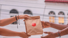 Muestra un primer plano de dos personas en el momento de la entrega de un pedido. Una mano sostiene y entrega una bolsa de papel kraft cerrada con una pegatina circular roja, mientras que la otra persona estira los brazos para recibirla en un entorno urbano.