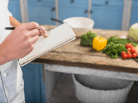 La imagen es un primer plano que se centra en las manos de una persona, probablemente un chef o cocinero, tomando notas en una cocina. La escena sugiere planificación o documentación de recetas.