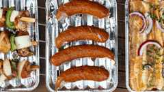 A imagem mostra uma vista aérea de três bandejas de alumínio descartáveis dispostas lado a lado sobre uma superfície de madeira escura, sugerindo preparo para churrasco ou grelhados.