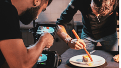À esquerda, um profissional (videomaker ou fotógrafo) com barba segura uma câmera equipada com um monitor externo. A tela exibe exatamente o ângulo do prato que está sendo capturado.