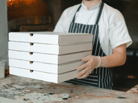 A imagem mostra um profissional de cozinha, com avental listrado e luvas de higiene, segurando uma pilha de caixas de pizza prontas para entrega. O cenário sugere uma operação focada exclusivamente em preparo e despacho de pedidos — sem atendimento ao cliente no local.
Essa é uma representação clássica do funcionamento de uma dark kitchen